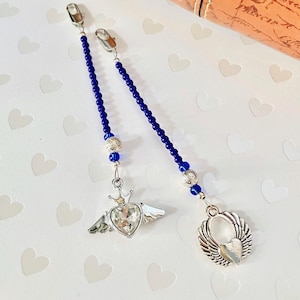 May include: Two silver-toned eyeglass chains with blue beaded strands. Each chain has a decorative charm: one with a heart-shaped crystal and crown with wings, and the other with a heart and wings. The chains are displayed on a white surface with heart-shaped patterns.
