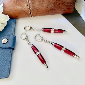 May include: Three red pen keychains with silver accents and a sparkling band. The pens are on a white surface next to a blue leather wallet and a brown leather bag. A white flower is in the background.