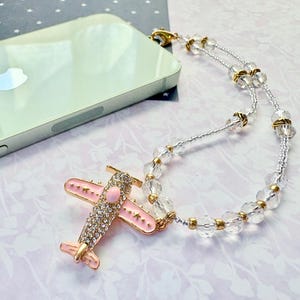 May include: A decorative phone charm featuring a pink airplane pendant adorned with rhinestones and gold accents. The charm is attached to a clear beaded chain with gold spacers. A silver phone rests in the background.