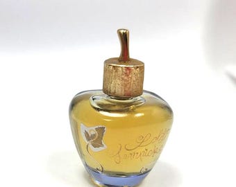 May include: A small, heart-shaped perfume bottle with a gold-colored cap and a blue base. The bottle is filled with a golden liquid and features the words "Lolita Lempicka" in elegant script, along with a floral design.