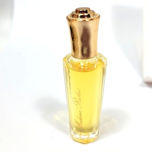 Op de afbeelding: Een kleine, heldere glazen parfumfles met een goudkleurige dop. De fles bevat een gele vloeistof en heeft de woorden "Madame Rochas" in gouden letters op de voorkant. De fles heeft een unieke, elegante vorm.