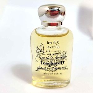 Könnte beinhalten: Eine kleine, klare Glasflasche von Cacharel Pour Homme Eau de Toilette. Die Flasche hat einen silbernen Verschluss und enthält eine hellgelbe Flüssigkeit. Das Etikett zeigt schwarze Schrift und das Cacharel-Logo.