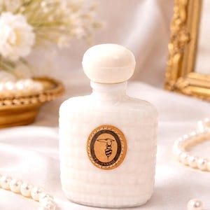 Peut inclure: Un flacon de parfum blanc avec une surface texturée et un bouchon rond blanc cassé. Une étiquette circulaire avec un logo est apposée sur le flacon. Le flacon est entouré de perles et d'un cadre doré décoratif.