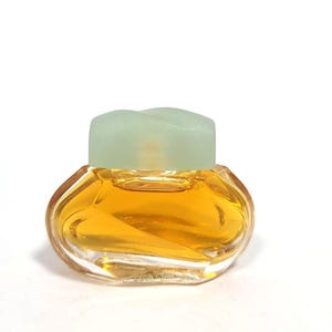 May include: A small, clear glass bottle of perfume with a light green cap. The bottle has a unique, curved shape and is filled with a golden liquid.