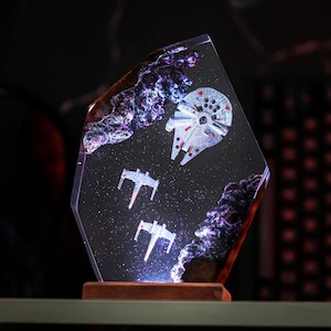 Könnte beinhalten: Klare, geometrische Kunstharzskulptur mit einer Weltraumszene, die einen Millennium Falken und zwei X-Wing-Sternjäger zeigt. Das Kunstwerk umfasst lila und weiße nebelartige Formationen und ist auf einem Holzsockel montiert.