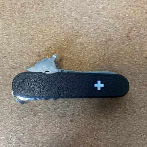 May include: A black multi-tool pocket knife featuring a textured handle with a white Swiss cross emblem. The tool is shown in a closed position with a metal bottle opener and screwdriver component partially extended from the top edge.