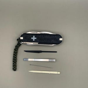 Puede incluir: Una multiherramienta negra y plateada con un emblema de cruz blanca, una regla y un cordón verde. Las herramientas adicionales incluyen un cuchillo negro, una pequeña herramienta plateada, un alfiler y una herramienta blanca.