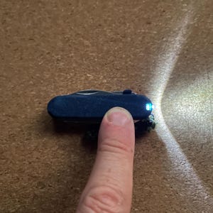 Op de afbeelding: Een donkerblauwe multitool met een kleine LED-zaklamp wordt getoond. De zaklamp is ingeschakeld en straalt een heldere witte straal uit. De tool wordt geactiveerd door een vinger. De achtergrond is een bruin, getextureerd oppervlak.
