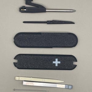 Op de afbeelding: Een assortiment zwarte en zilveren multi-tool componenten, waaronder een mes, een schroevendraaier en een handvat met textuur en een wit kruis embleem. De gereedschappen zijn gerangschikt op een lichtgrijze ondergrond.