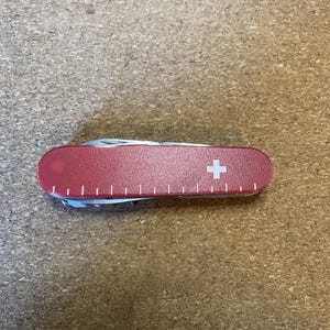 Op de afbeelding: Een rood Zwitsers zakmes met een wit kruis-embleem en een liniaalschaal geprint langs de onderrand van het handvat. De multitool is gesloten en toont de gladde, afgeronde behuizing en de metalen onderdelen die aan de randen zichtbaar zijn.