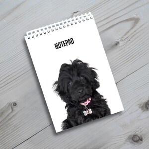 May include: A white spiral-bound notepad featuring the word NOTEPAD in black text at the top and a photograph of a fluffy black puppy wearing a pink collar with a bone-shaped silver tag centred on the front cover.