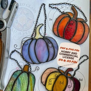 May include: Four stained glass pumpkin suncatchers in purple, orange, green, and yellow-pink, each with a black chain. They are displayed on a paper pattern titled Simple Pumpkins Suncatcher Pattern by Ad Astra Stained Glass with PDF & PNG File text.