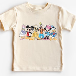 May include: Cream-colored t-shirt featuring a colorful graphic of Disney characters, including Mickey Mouse, Minnie Mouse, Donald Duck, and Goofy. The design has a watercolor style with black outlines and bright colors. The shirt is made of soft material.