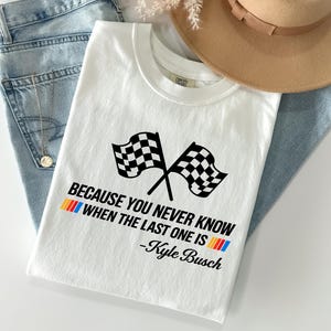 May include: White t-shirt with a crew neck featuring two crossed checkered flags and the text "BECAUSE YOU NEVER KNOW WHEN THE LAST ONE IS -Kyle Busch" in black, red, yellow, and blue. The shirt is laid on top of blue jeans and a tan hat.