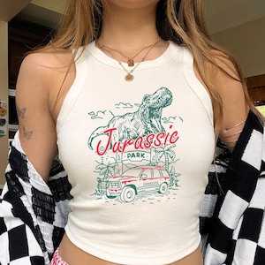 Può includere: Canotta color crema con grafica "Jurassic Park" verde e rossa raffigurante un T-Rex, una jeep rossa e palme. La canotta è abbinata a pantaloncini a quadri rossi e bianchi. Un cardigan a quadri bianchi e neri è drappeggiato sulle spalle.