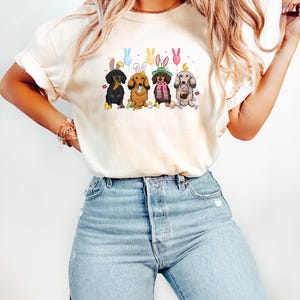 May include: Cream-colored t-shirt featuring a graphic of five dachshund dogs dressed for Easter. Each dog wears bunny ears and holds Easter eggs or balloons. The dogs are in a row, with colorful accents.