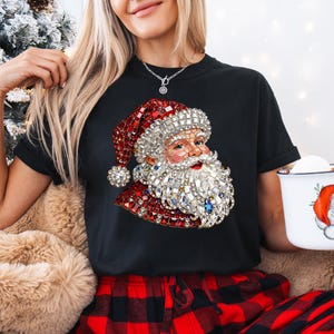 Może przedstawiać: Czarny t-shirt z grafiką Świętego Mikołaja, z czerwonym kapeluszem i brodą z błyszczącymi zdobieniami. Widoczny jest r&oacute;wnież biały kubek z ilustracją Świętego Mikołaja, a także czerwono-czarne spodnie w kratę.