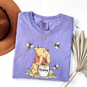 May include: A purple t-shirt with a cartoon image of Winnie the Pooh holding a pot of honey. The shirt has the text "Hunny" on the pot of honey. There are bees flying around Winnie the Pooh.