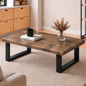 May include: A rectangular coffee table featuring a thick, dark brown stained wooden top with visible wood grain and knots, supported by two sturdy, matte black metal U-shaped legs. The surface displays a black mug and a book titled KINFOLK.