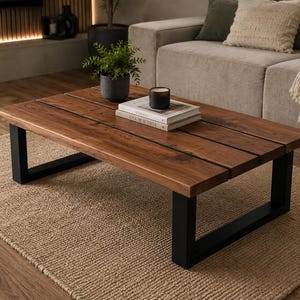 May include: A rectangular coffee table featuring a thick, natural-toned wooden top composed of three parallel planks with visible grain, supported by two sturdy, matte black U-shaped metal legs. A small potted plant and two stacked books sit on top.