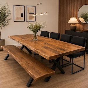 Op de afbeelding: Een rechthoekige eettafel en bijpassende bank met natuurlijke houten bladen met zichtbare nerven, ondersteund door zwarte X-vormige metalen poten. Vier zwarte gestoffeerde stoelen zijn langs de zijkant van de tafel geplaatst.