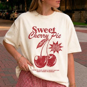 May include: A cream-colored t-shirt featuring a red graphic of two cherries with the text Sweet Cherry Pie, Oh Sh*t Graham You Know This One?, and Tastes So Good, Make A Grown Man Cry Sweet Cherry Pie-eeeeee, paired with red and white gingham shorts.