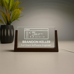 Op de afbeelding: Een rechthoekig bureaubord met een donkere houten basis en een heldere acryl voorkant. Het acryl toont een witte afbeelding van een codeervenster en de naam "BRANDON KELLER, Full Stack Software Engineer."