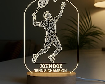 Op de afbeelding: Een verlicht nachtlampje met een tennis thema. Het ontwerp toont een tennisser in actie, met een racket en bal. De basis is van hout en de tekst "JOHN DOE TENNIS CHAMPION" is zichtbaar. Het licht heeft een warme witte kleur.