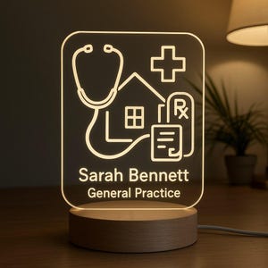 May include: A light-up acrylic sign with a wooden base. The sign displays a stethoscope, house, cross, and prescription symbols. The text reads "Sarah Bennett General Practice". The light emits a warm white glow.