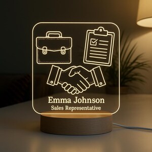 May include: A personalized LED desk lamp with a wooden base. The lamp features a white outline design of a briefcase, clipboard, and handshake. The name "Emma Johnson" and "Sales Representative" are printed below the images.