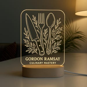 May include: A rectangular LED lamp featuring a knife, fork, and spoon design with leafy accents. The text "GORDON RAMSAY CULINARY MASTERY" is displayed. The lamp sits on a wooden base and is plugged in.