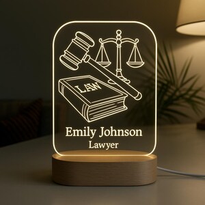 Peut inclure: Lampe de bureau en acrylique &eacute;clair&eacute;e avec une base en bois. La lampe repr&eacute;sente un maillet, une balance de la justice et un livre avec le mot "LAW" grav&eacute;. Le nom "Emily Johnson" et le titre "Lawyer" sont &eacute;galement affich&eacute;s.