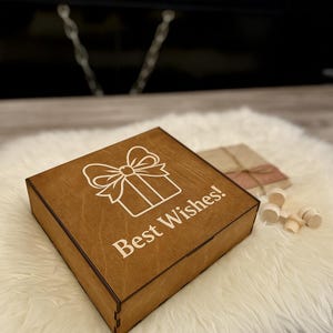 Peut inclure: Une bo&icirc;te cadeau carr&eacute;e en bois avec un motif de cadeau et de n&oelig;ud grav&eacute; au laser, et les mots "Best Wishes!" en blanc. La bo&icirc;te est sur une surface blanche et moelleuse, avec d'autres cadeaux et des pi&egrave;ces en bois en arri&egrave;re-plan.