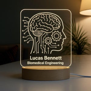Può includere: Una luce notturna a LED personalizzata con un design a cervello e circuito stampato. La luce è incisa con il nome "Lucas Bennett" e il titolo "Biomedical Engineering". Si trova su una base di legno ed è collegata a una fonte di alimentazione.