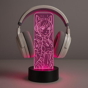 Op de afbeelding: Een neonroze LED-lamp met een manga-stijl karakter met kattenoren, geplaatst op een zwarte basis. Een witte en grijze koptelefoon is rond het licht geplaatst, wat een gaming-thema creëert. Een decoratief element.
