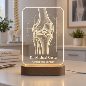 Könnte beinhalten: Eine rechteckige LED-Schreibtischlampe aus Acryl mit einer geätzten anatomischen Darstellung eines menschlichen Kniegelenks. Der Sockel besteht aus glattem braunem Holz. Der Text lautet Dr. Michael Carter Orthopedic Surgery in weißer Schrift.