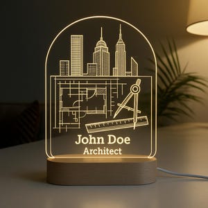 Peut inclure: Une veilleuse architecturale personnalis&eacute;e avec un design de paysage urbain, un plan et des outils de dessin. La lumi&egrave;re est grav&eacute;e du nom "John Doe" et du titre "Architecte". La base est en bois.