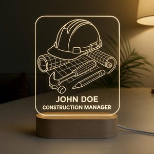 Peut inclure: Lampe de chevet personnalis&eacute;e sur le th&egrave;me de la construction. Elle pr&eacute;sente un casque de chantier, des plans et des outils, avec le nom "JOHN DOE" et le titre "CONSTRUCTION MANAGER". La lampe est pos&eacute;e sur un socle en bois.