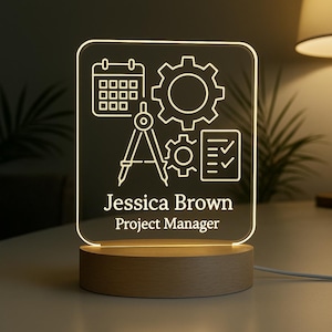 May include: A personalized LED desk lamp with a wooden base. The clear acrylic panel features white line art of engineering icons and the text "Jessica Brown, Project Manager."