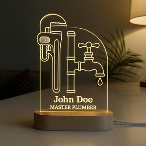 May include: A lighted acrylic plumber's night light with a wooden base. The design features a wrench, pipes, and a faucet. The name "John Doe" and the title "MASTER PLUMBER" are displayed in white text.