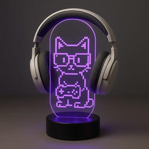 以下が含まれることがあります： メガネをかけたピクセル化された猫がゲームコントローラーを持っている紫色のLEDナイトライト。ライトは黒いベースにあり、両側に灰色のヘッドフォンがあります。猫のデザインは明るく光っています。
