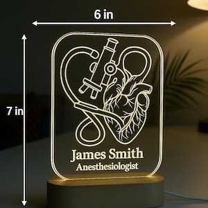 Peut inclure: Lampe LED rectangulaire avec une base en bois, pr&eacute;sentant un design m&eacute;dical. Le panneau lumineux affiche un microscope, un c&oelig;ur et le nom "James Smith, Anesth&eacute;siste". La lampe mesure 15 cm de large et 18 cm de haut.