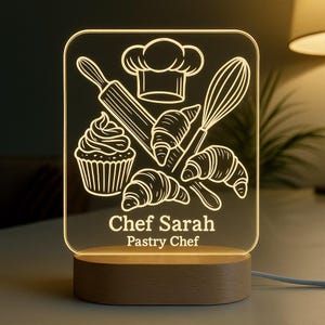 Könnte beinhalten: Ein personalisiertes Nachtlicht mit Holzsockel. Die transparente Acrylplatte zeigt eine Kochmütze, ein Nudelholz, einen Schneebesen, Croissants und einen Cupcake. Der Text lautet "Chef Sarah, Konditor."