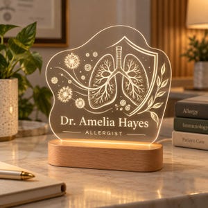 Puede incluir: Un letrero de escritorio de acrílico iluminado con base de madera, grabado con una ilustración anatómica de pulmones, semillas de diente de león y el texto Dr. Amelia Hayes ALLERGIST. El panel transparente brilla con luz cálida.