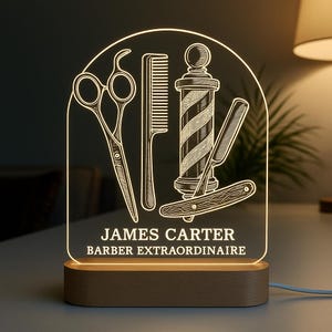 Pode incluir: Uma luz noturna LED personalizada para barbearia. A luz apresenta um design de poste de barbeiro, tesoura, pente e navalha. O texto "JAMES CARTER BARBER EXTRAORDINAIRE" é exibido na luz. A base é de madeira.