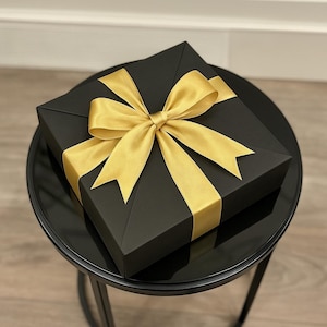 Peut inclure: Une bo&icirc;te cadeau carr&eacute;e noire avec un ruban de satin dor&eacute; nou&eacute; en n&oelig;ud. La bo&icirc;te est pos&eacute;e sur une petite table ronde noire. Le n&oelig;ud est centr&eacute; sur le dessus de la bo&icirc;te.