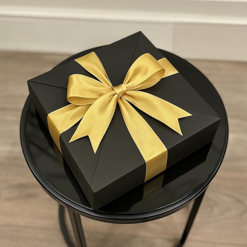 Peut inclure: Une bo&icirc;te cadeau carr&eacute;e noire avec un ruban de satin dor&eacute; nou&eacute; en n&oelig;ud. La bo&icirc;te est pos&eacute;e sur une petite table ronde noire. Le n&oelig;ud est centr&eacute; sur le dessus de la bo&icirc;te.
