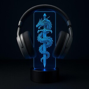 Könnte beinhalten: Schwarzer Over-Ear-Kopfhörer, der ein leuchtendes blaues LED-Licht umrahmt. Das Licht zeigt eine detaillierte Illustration eines Drachen, der sich um ein Schwert windet. Der Sockel des Lichts ist schwarz, und der Hintergrund ist dunkel.