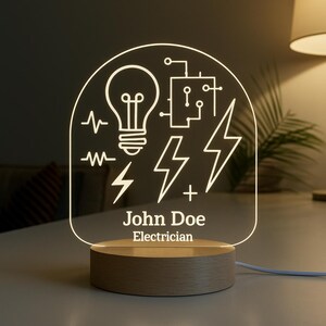 May include: A personalized LED night light with a wooden base. The clear acrylic panel features an electrician-themed design with a lightbulb, lightning bolts, and the text "John Doe Electrician". The light is on, illuminating the design.