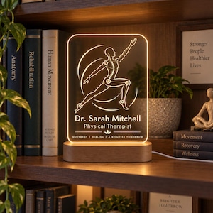 Può includere: Un'insegna luminosa rettangolare in acrilico con base in legno, caratterizzata da una figura umana incisa in posa dinamica, il testo Dr. Sarah Mitchell Physical Therapist e le parole MOVEMENT HEALING A BRIGHTER TOMORROW con una piccola icona a foglia.